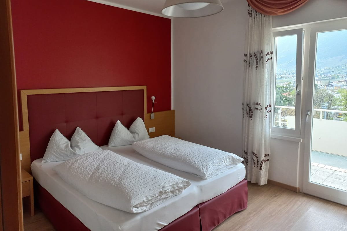 A double bed with white bedding, a wooden headboard, and a view of a balcony with a cityscape.