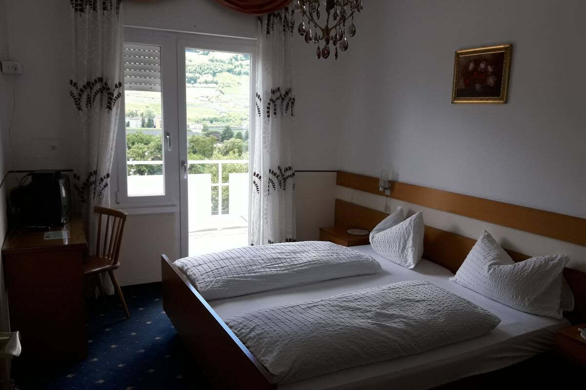 A hotel room with a bed, a chair, a desk with a television, a window with curtains, and a picture on the wall.