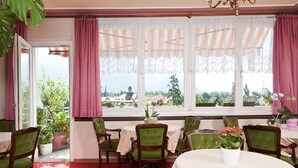 A dining area with a table and chairs, large windows with curtains, and potted plants. - Hotel Garni Lux (Merano)