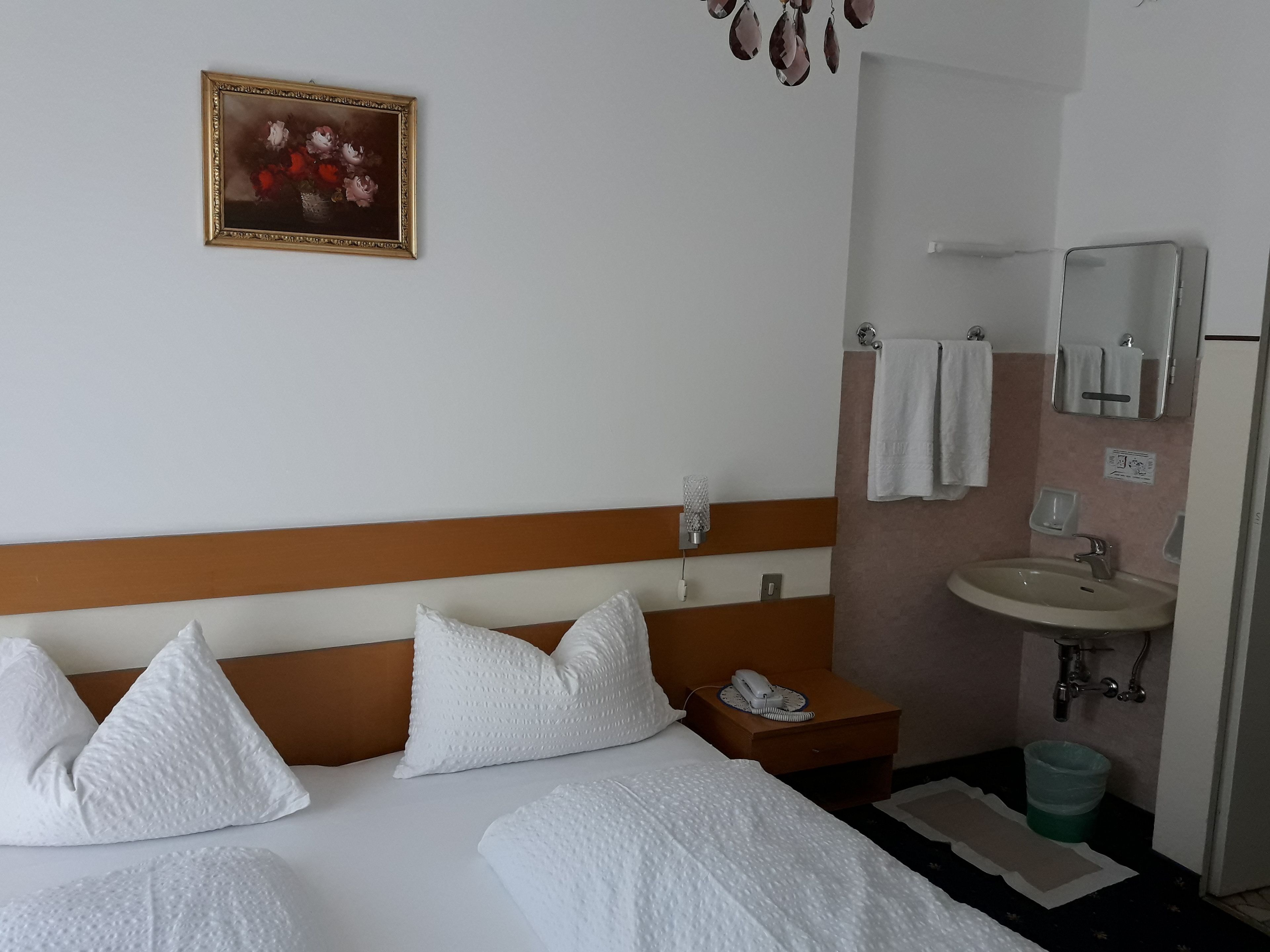 A hotel room with a bed, pillows, a bedside table with a phone, a wall-mounted painting, a bathroom with a sink and mirror, and a trash bin.