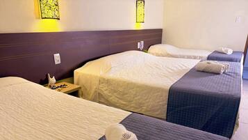 Ein Hotelzimmer mit drei Betten, jedes mit einer anderen farbigen Bettdecke. Es gibt Wandleuchten mit gelbem Licht, einen kleinen Tisch mit einer Uhr und eine weiße Katzenfigur auf dem Nachttisch.