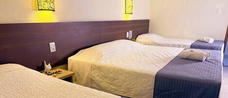 A hotel room with three beds, each with a different colored bedspread. There are wall sconces with yellow light, a small table with a clock, and a white cat figurine on the nightstand.