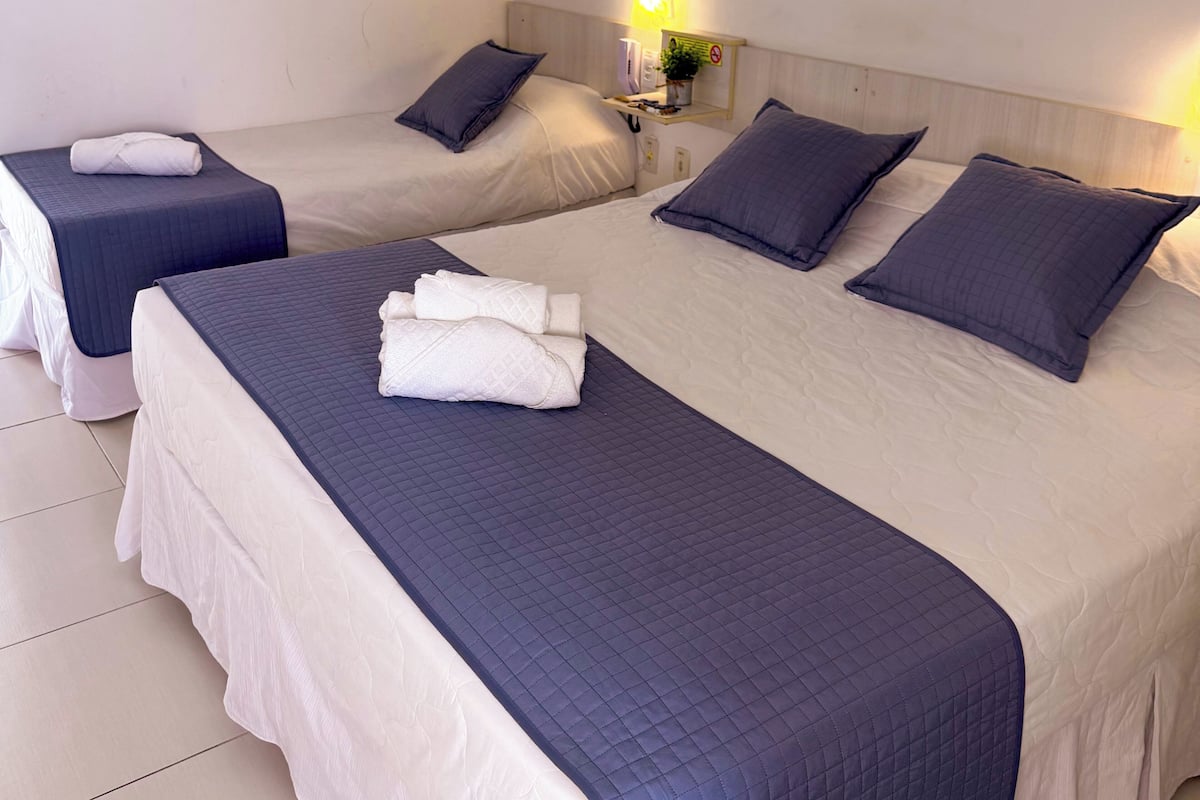 Habitación de hotel con dos camas, cada una con una colcha acolchada azul y almohadas. Hay una mesita de noche pequeña con una lámpara y un cartel, y una planta en maceta en la pared.