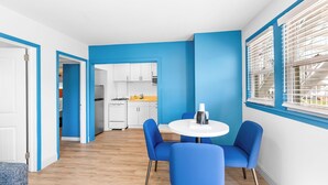 A modern kitchen with white cabinets and a stove, a dining area with blue chairs and a white table, and a bright blue accent wall. - Buttercup Suites (Wildwood Crest)