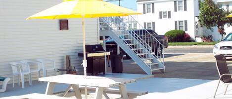 Une table de pique-nique avec un parasol jaune, un barbecue et une poubelle, devant un bâtiment blanc doté d’un escalier.