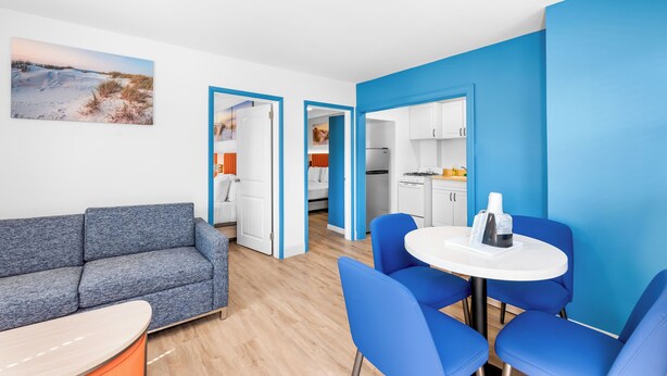 A modern living room with a grey sofa, blue chairs, and a white table. There is a kitchen area visible through an open doorway. - Buttercup Suites (Wildwood Crest)