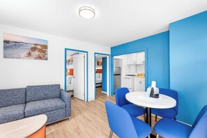 A modern living room with a grey sofa, blue chairs, and a white table. There is a kitchen area visible through an open doorway. - Buttercup Suites (Wildwood Crest)