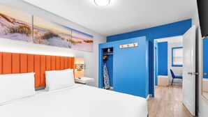 A modern bedroom with a large bed, a nightstand, a wall-mounted shelf, and a door leading to another room. - Buttercup Suites (Wildwood Crest)