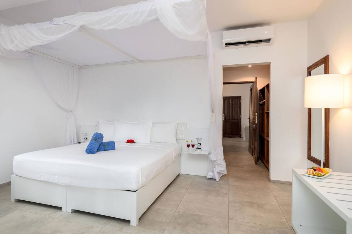 A hotel room with a bed, white curtains, a bedside table with a lamp, an air conditioning unit, and a hallway leading to another room.