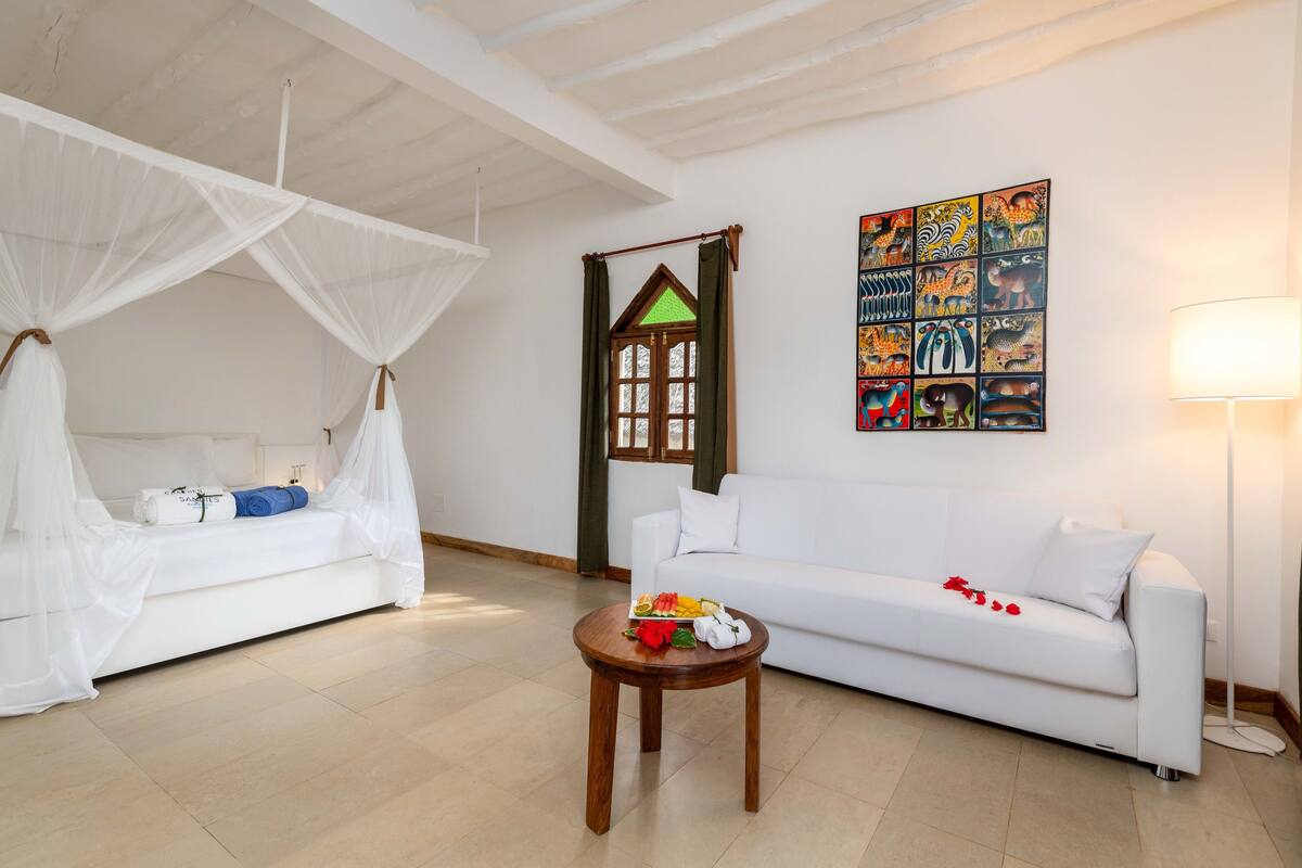 A room with a bed under a mosquito net, a white sofa, a small wooden table with a fruit bowl, and a wall with colorful animal paintings.