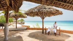 Un restaurante frente a la playa con sombrillas de paja, suelo de piedra y vistas al océano.