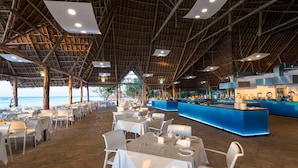 Un restaurante con techo de paja, mesas blancas y una zona de bar azul.
