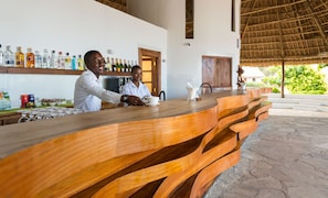 A bar area with a wooden counter, bottles of beverages, and a view of greenery outside.