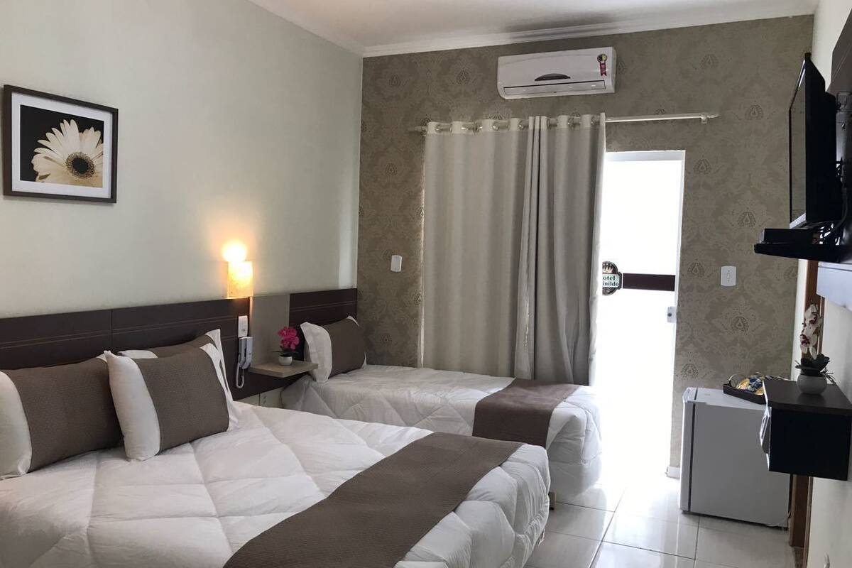 A hotel room with two beds, a nightstand with a lamp, an air conditioning unit, a small refrigerator, and a television on a stand.