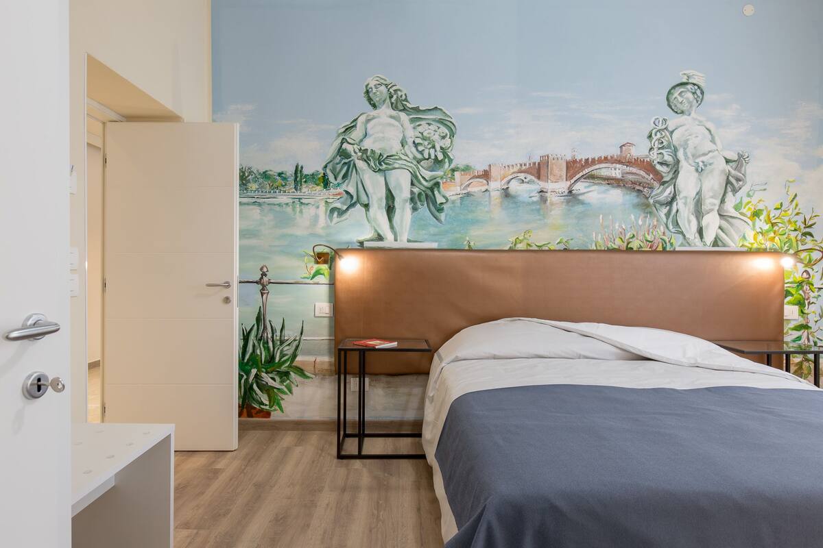 A bedroom with a mural of a bridge and figures, a bed, a bedside table, and a door.