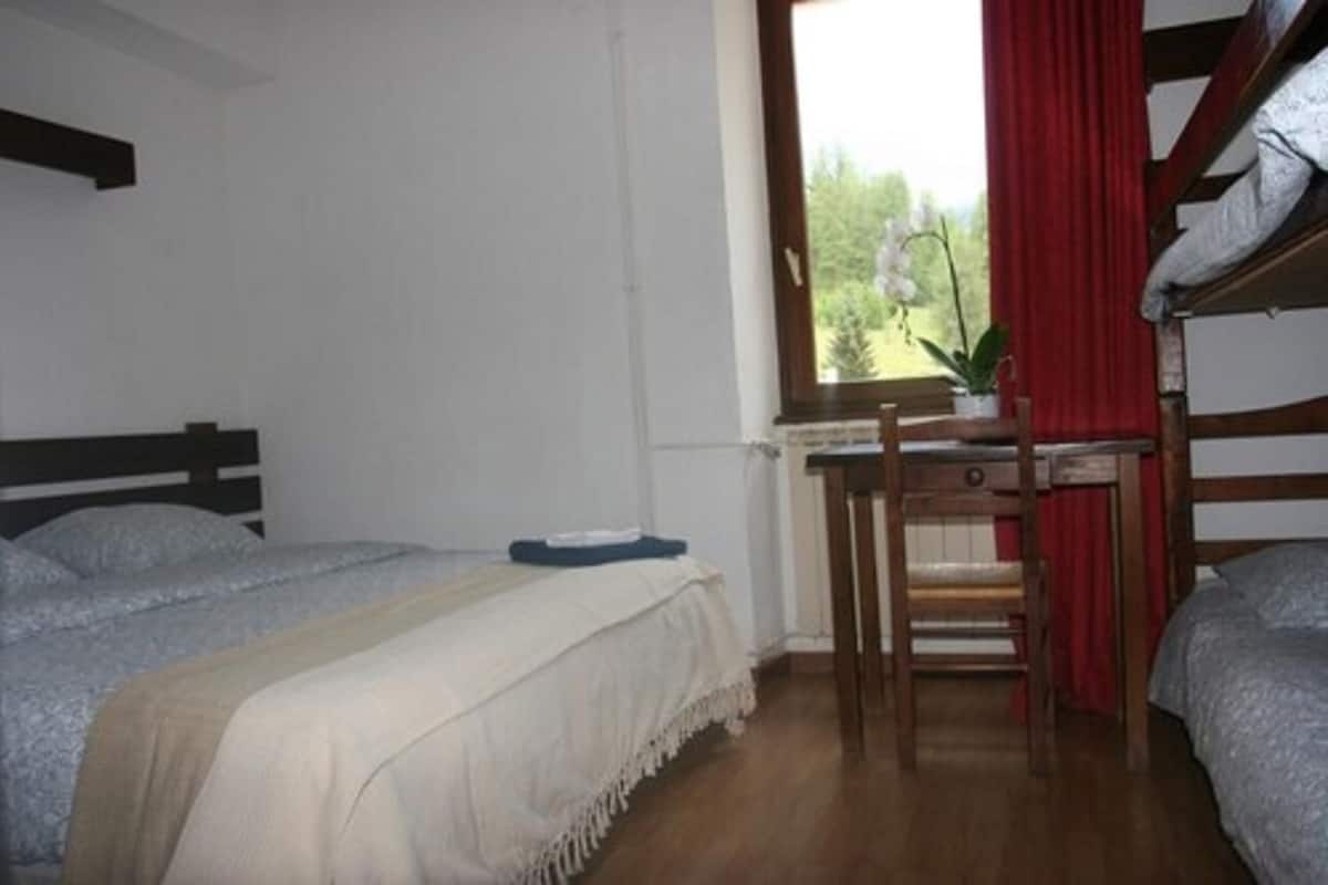 Een studentenkamer met twee bedden, een bureau, een stoel en een raam met uitzicht op groen.