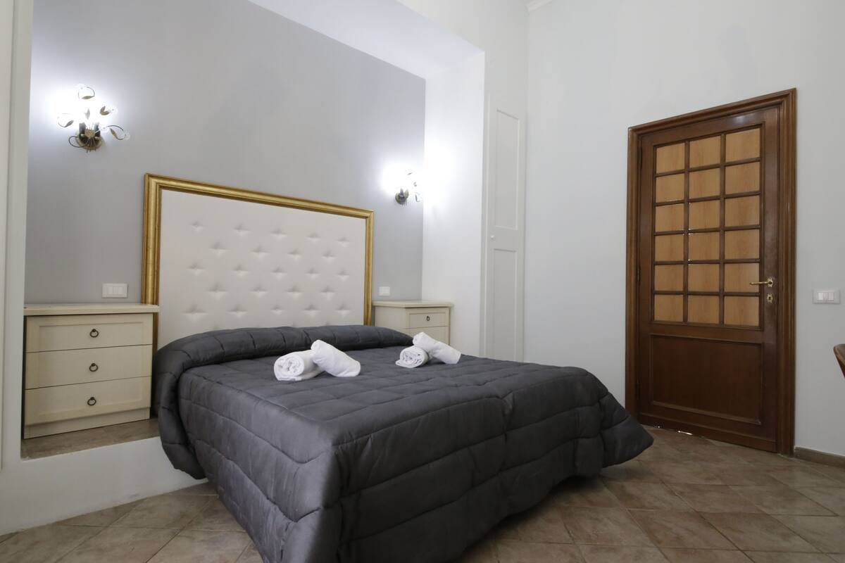 A bedroom with a large bed, two bedside tables, a wooden door, and a wall-mounted light fixture.
