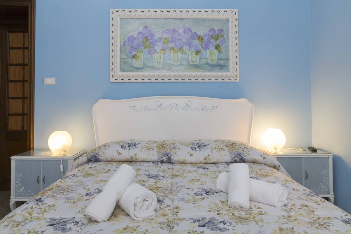 A bedroom with a floral bedspread, two bedside lamps, and a painting of purple flowers on the wall.