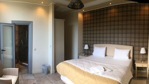 A hotel room with a bed, a chair, a nightstand, a lamp, a trash bin, and a shower area. - Akraion (Penteli)