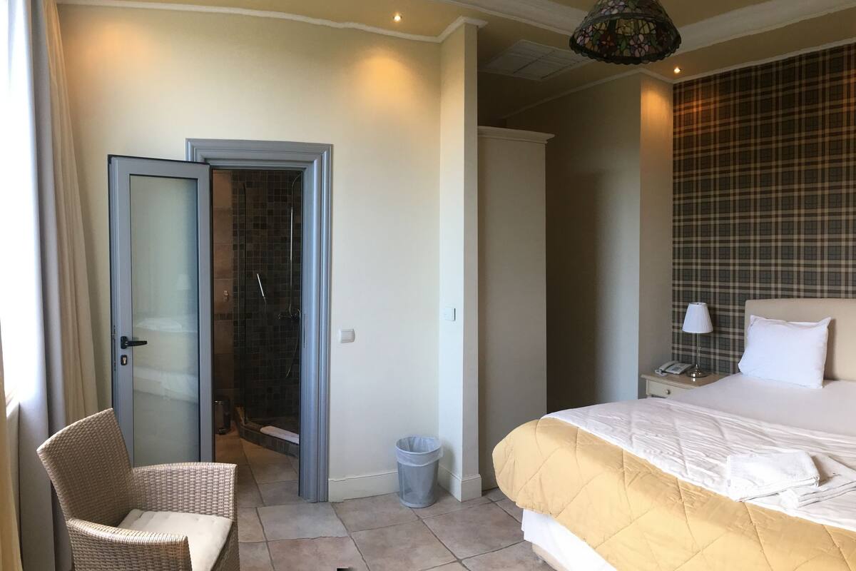A hotel room with a bed, a chair, a nightstand, a lamp, a trash bin, and a shower area.