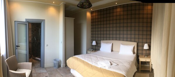 A hotel room with a bed, a chair, a nightstand, a lamp, a trash bin, and a shower area.