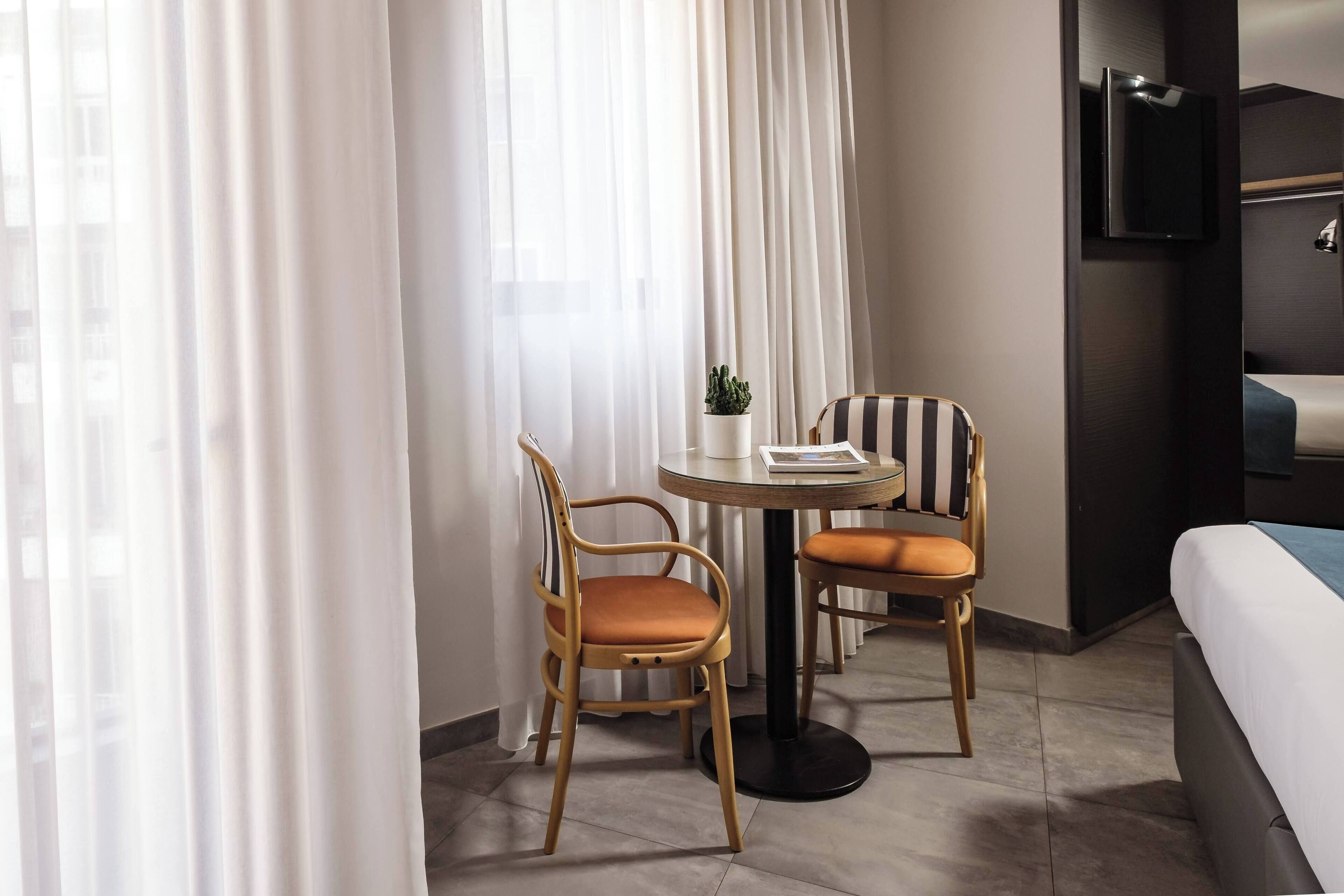 A hotel room with a bed, a TV, a small table with two chairs, and a window with curtains.