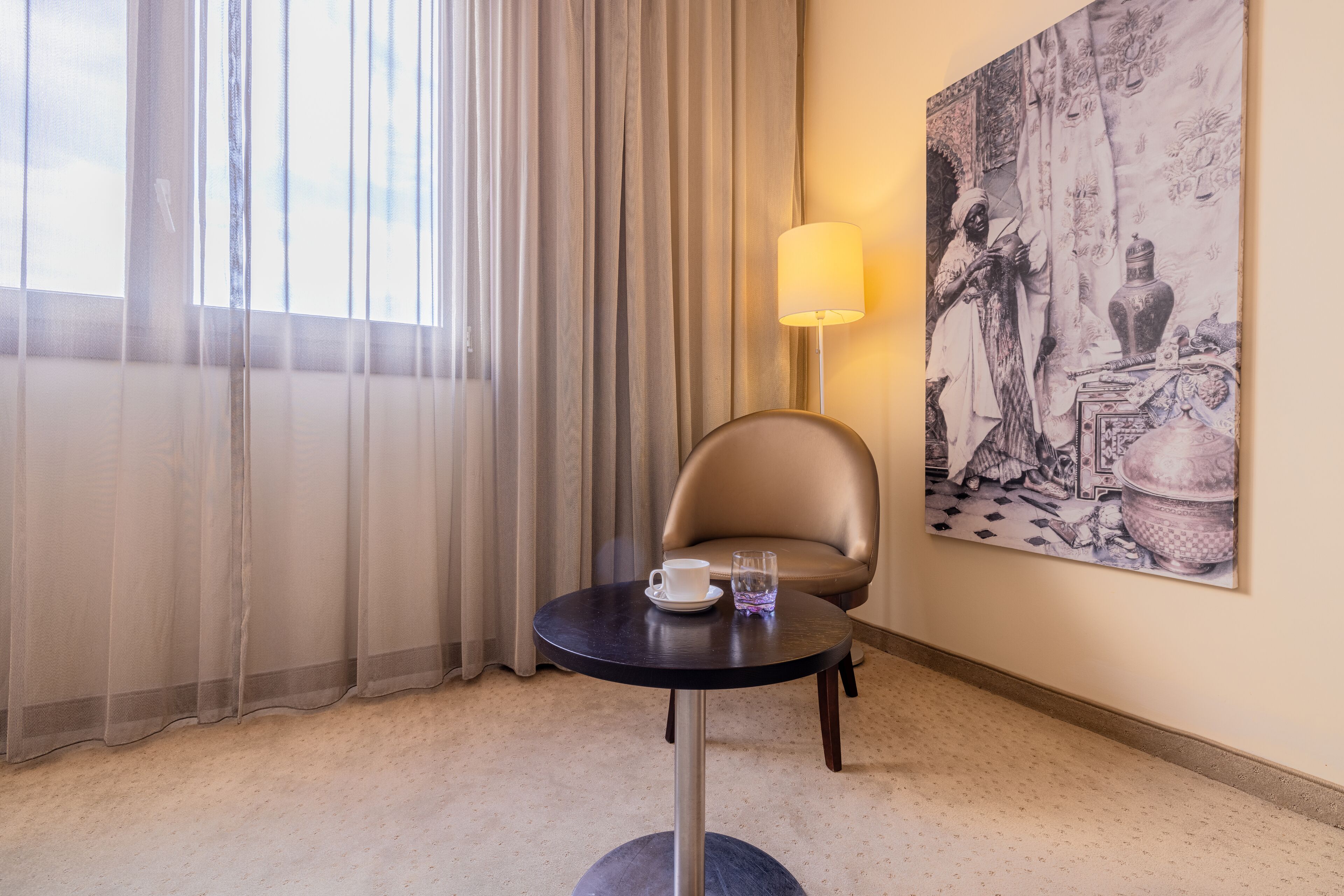 Ett hotellrum med ett runt bord, en stol, en lampa, en tavla på väggen och ett fönster med gardiner.