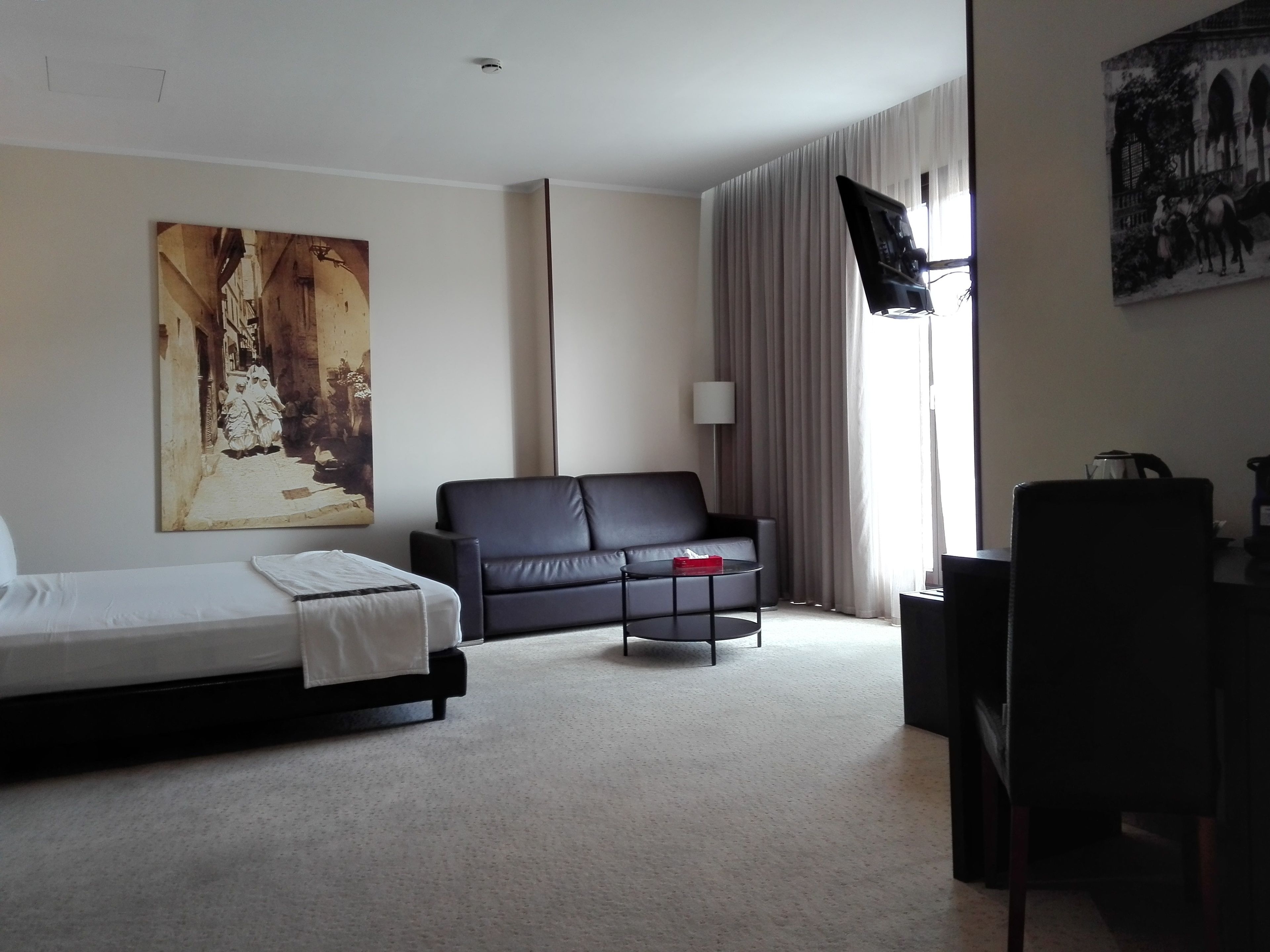 A hotel room with a bed, a black leather sofa, a small table, a desk, and a large painting on the wall.