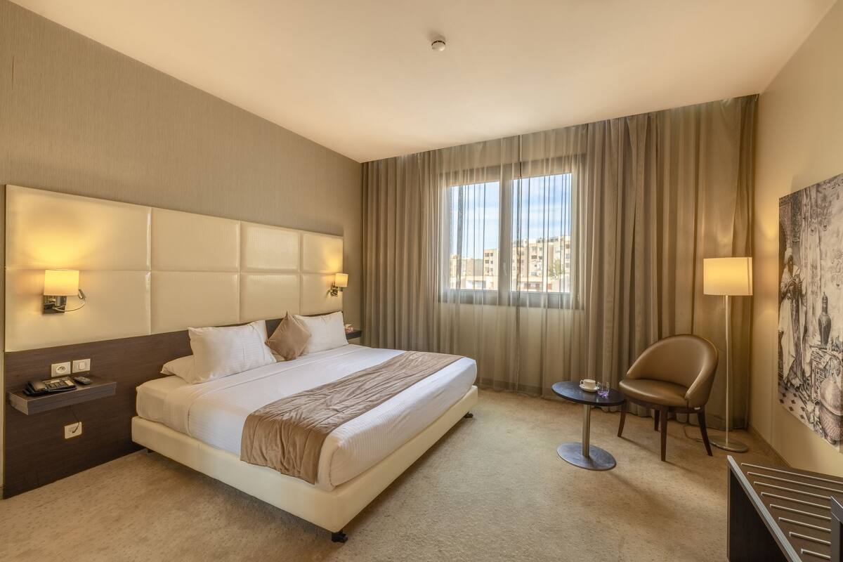 A hotel room with a large bed, bedside lamps, a small table, a chair, and a large window with curtains.
