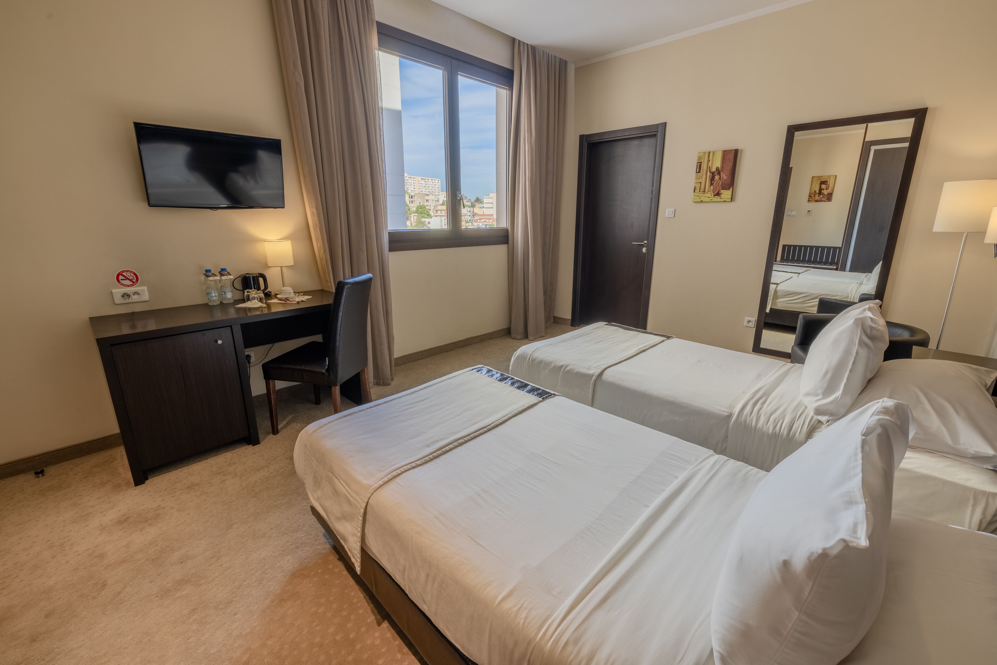 A hotel room with two beds, a desk, a chair, a TV, a window with curtains, and a door.