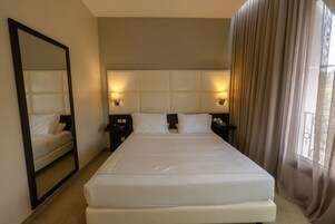 Habitación de hotel con una cama grande, cabecera con luces, un espejo y una ventana con cortinas.
