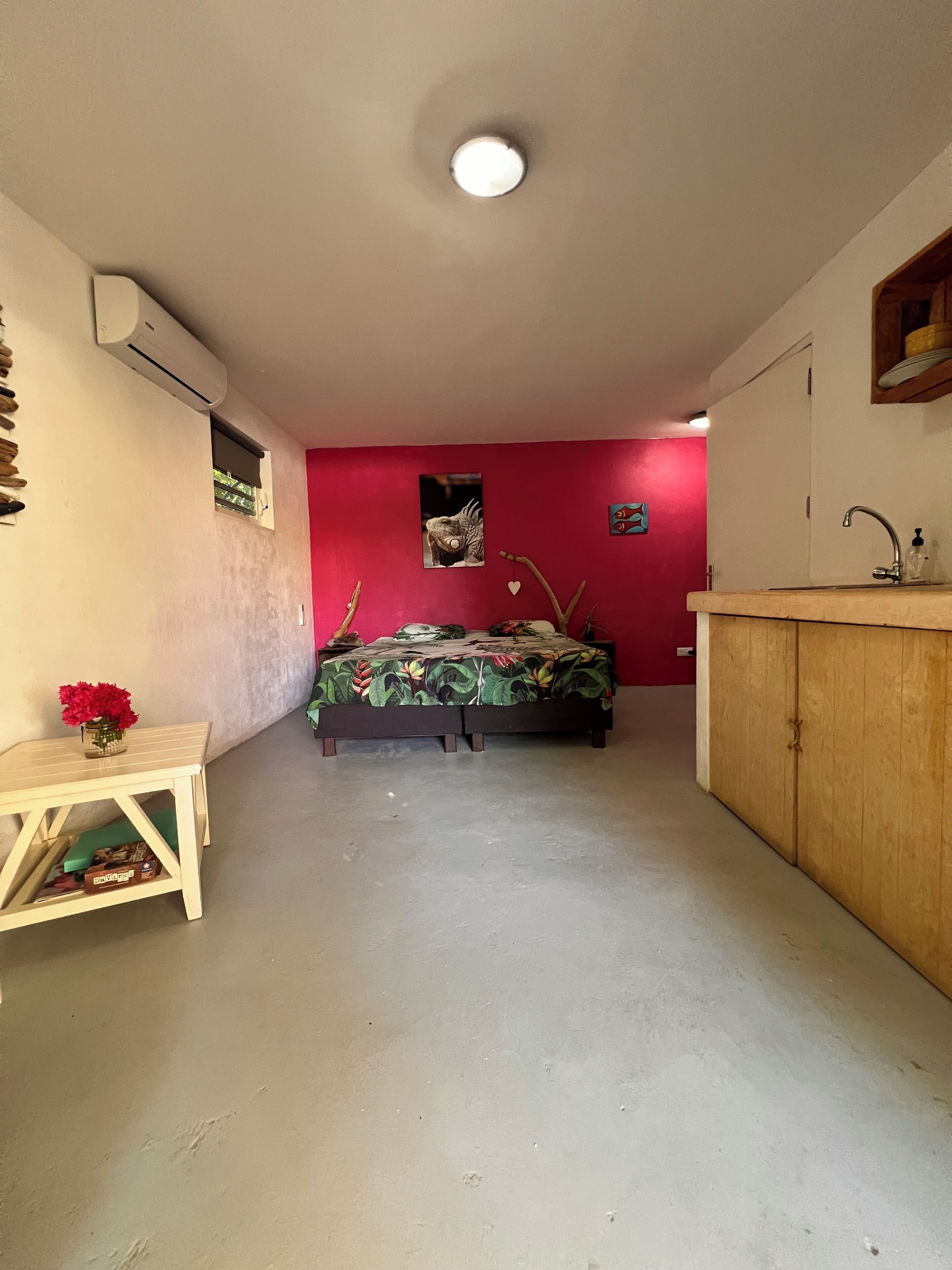 A room with a bed, a small table with a vase, a red wall, a wooden cabinet, and an air conditioning unit.