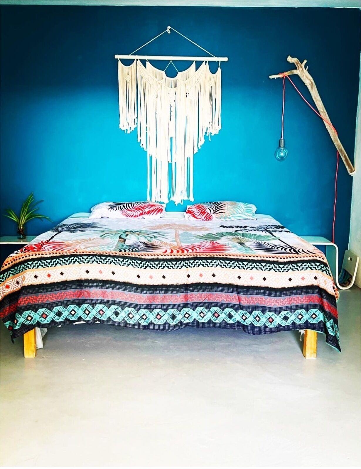 A bed with a patterned bedspread, a macrame wall hanging, and a hanging light fixture.