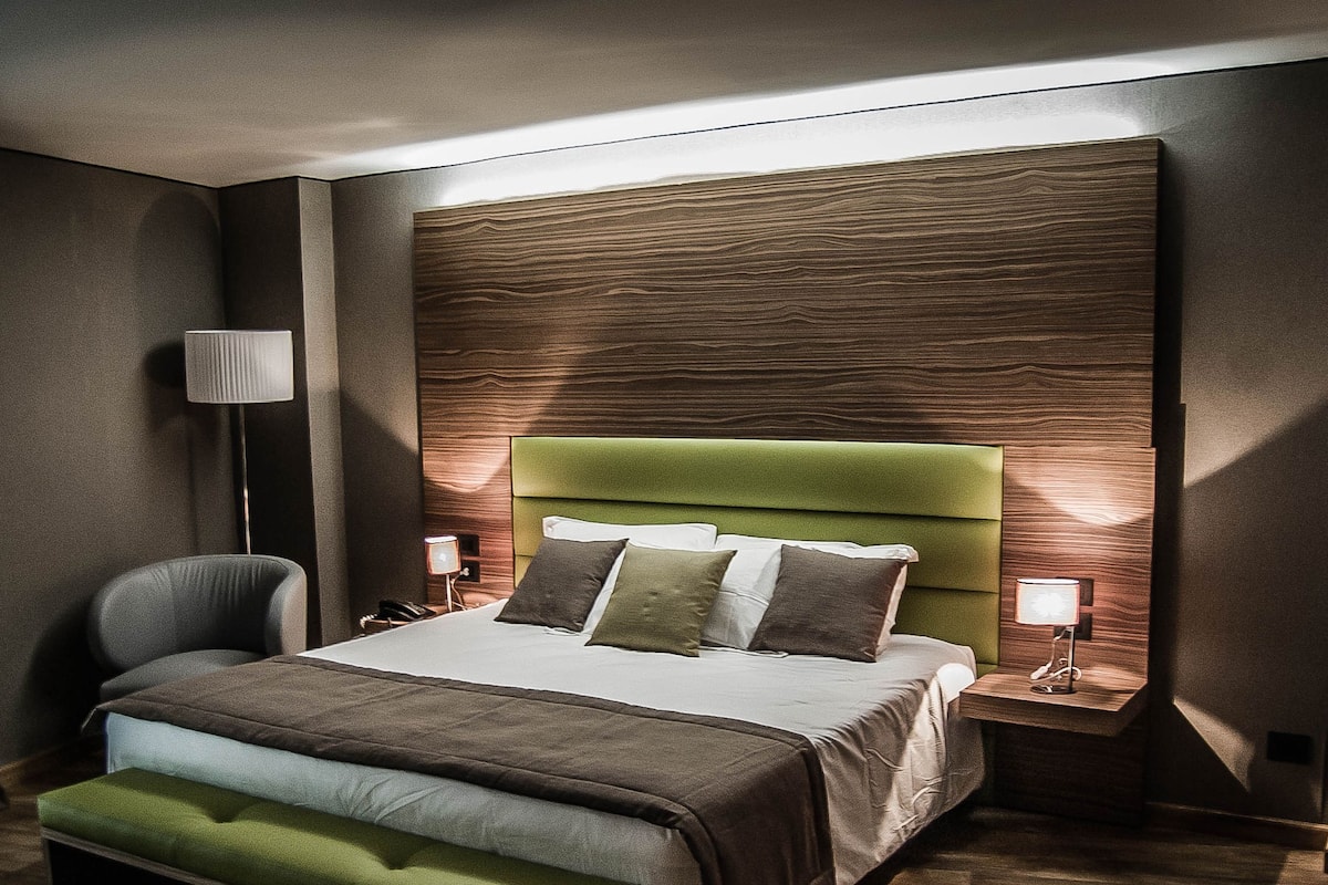Moderni hotellihuone, jossa on suuri sänky, vihreäverhoiltu pääty, yöpöytä lampun kanssa ja tuoli.