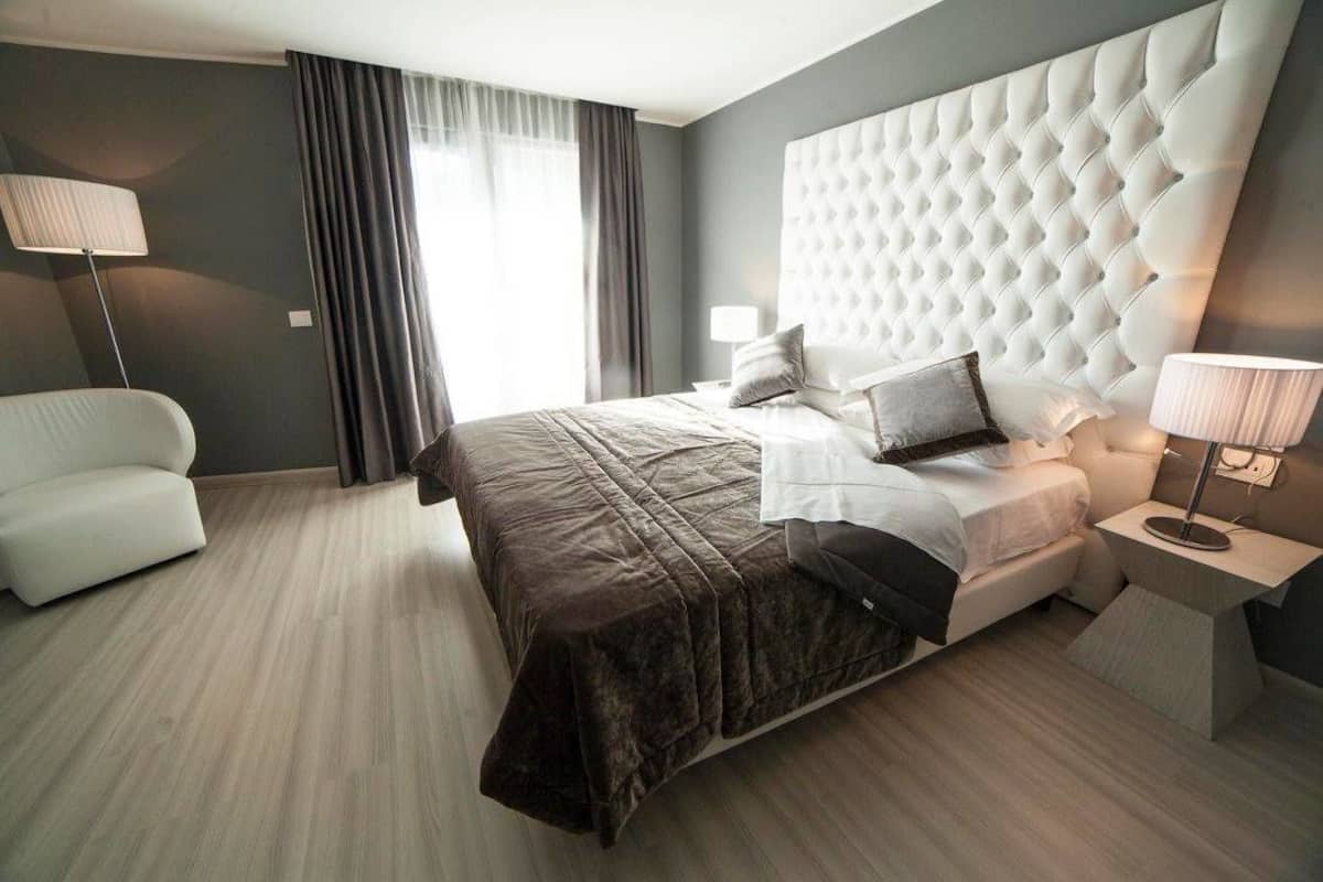 Moderni hotellihuone, jossa on suuri sänky, valkoinen, verhoiltu pääty, yöpöytä lampun kanssa ja valkoinen nojatuoli.