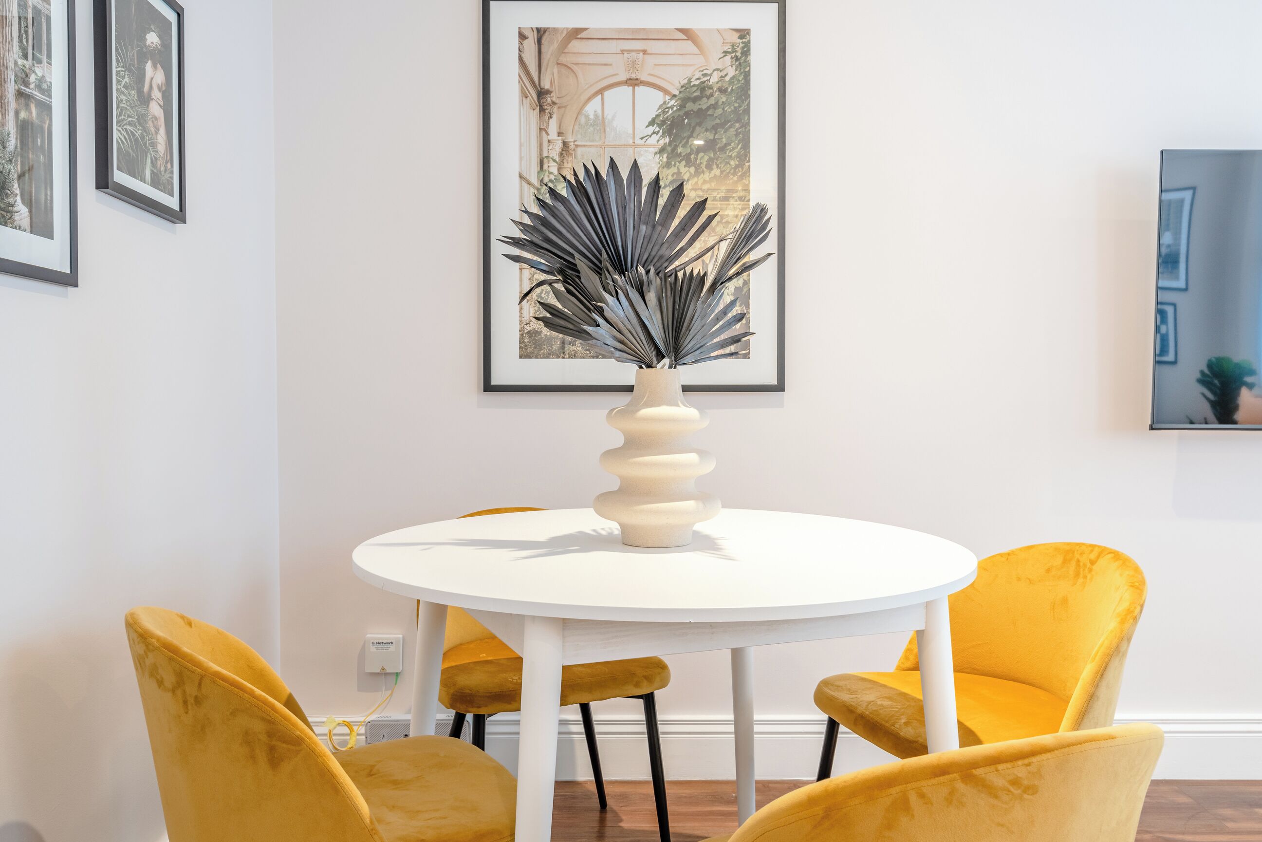 A round white table with a vase of dark foliage, surrounded by four yellow chairs.