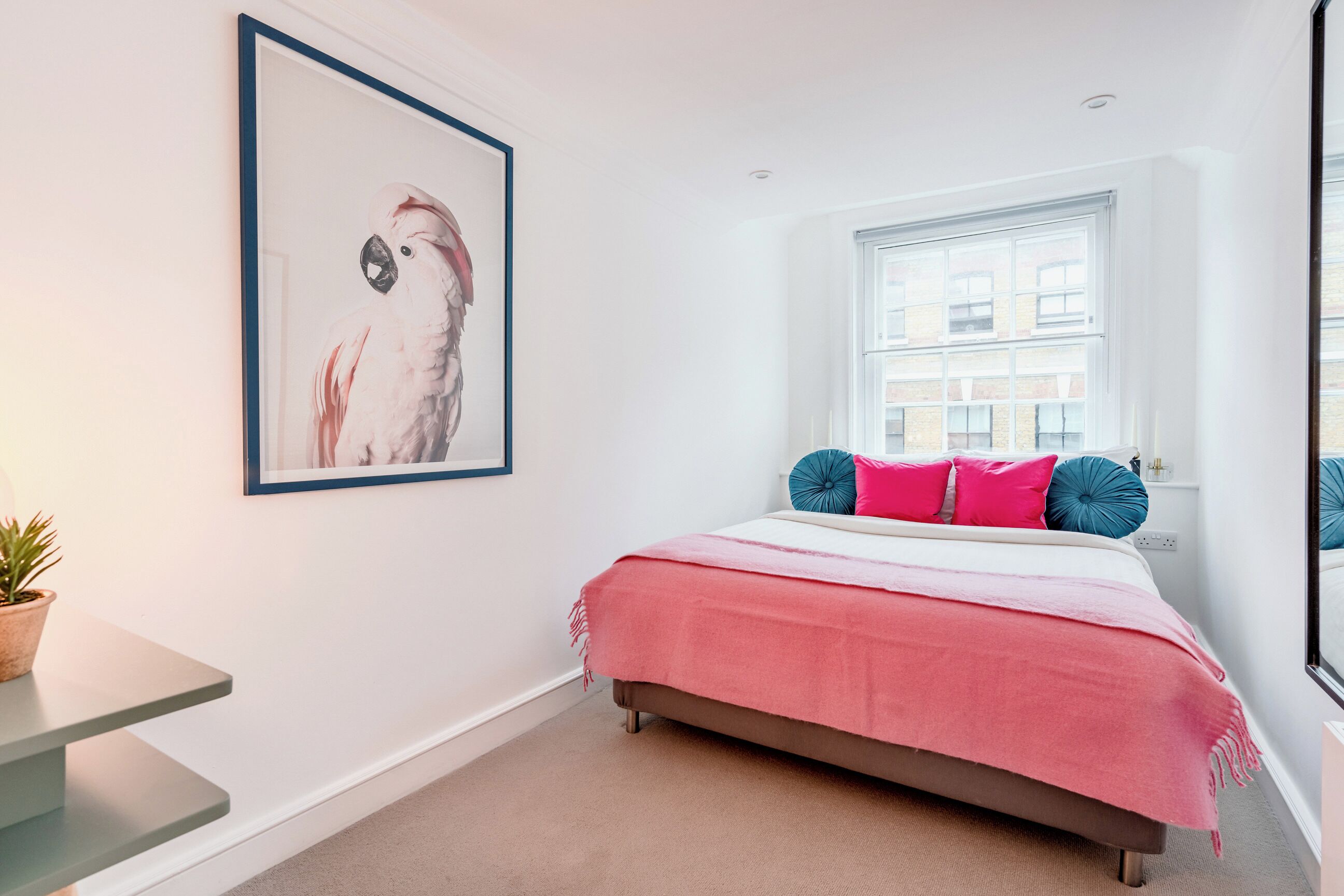 A bedroom with a bed, a framed picture on the wall, a window, and a small table with a plant.