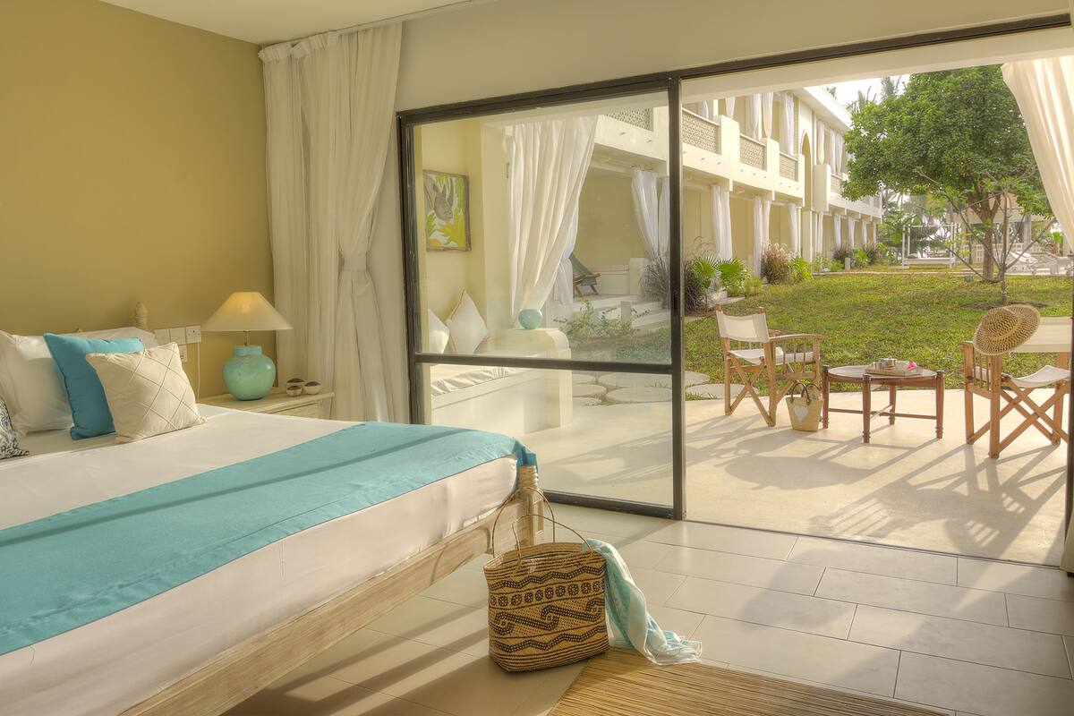Habitación de hotel con cama, mesita de noche, lámpara, cesta de mimbre y vistas al área de estar exterior.