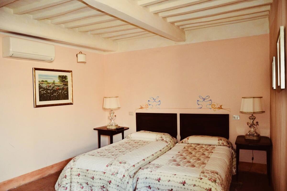 A bedroom with two beds, a nightstand with a lamp, a wall-mounted air conditioner, and a framed picture.