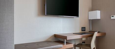 Una habitación de hotel moderna con un televisor de pantalla plana montado en la pared, un escritorio de madera con una silla, un banco y un pequeño cesto de basura.