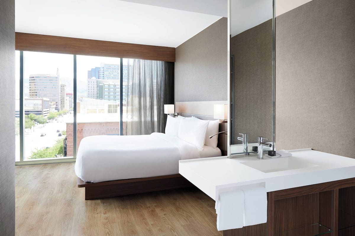 Une chambre d’hôtel moderne dotée d’un grand lit, d’une salle de bain blanche avec un lavabo et offrant une vue sur la ville grâce à de grandes fenêtres.
