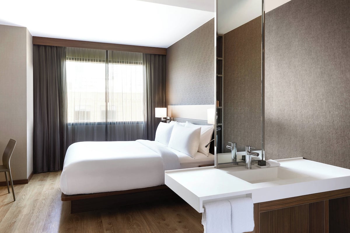 Une chambre d’hôtel moderne dotée d’un grand lit, d’un bureau intégré, d’un grand miroir et d’une salle de bain avec un lavabo et un meuble-lavabo blancs.