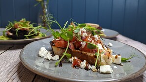 A plate of food with a slice of bread topped with tomatoes, cheese, and greens, and a vase with flowers in the background. - The Coach House Inn (Chester)