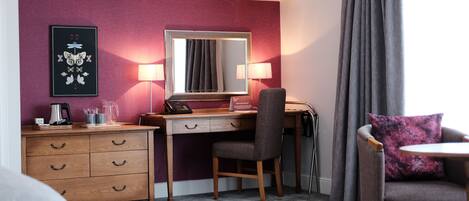 Habitación de hotel con escritorio de madera, una silla, cama, lámpara de escritorio, espejo, cuadro enmarcado y ventana con cortinas.