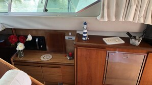 Een bescheiden bootinterieur met een houten bureau, een kleine koelkast, een radio en een raam met uitzicht op het water.