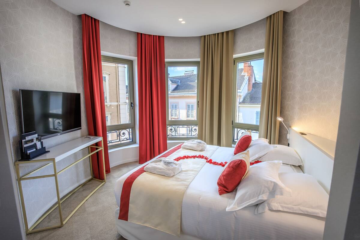 A hotel room with a bed, a TV, a desk, and large windows with curtains.