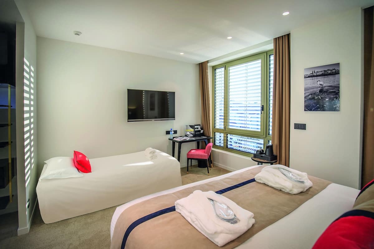 A hotel room with two beds, a desk, a TV, and a window with blinds.