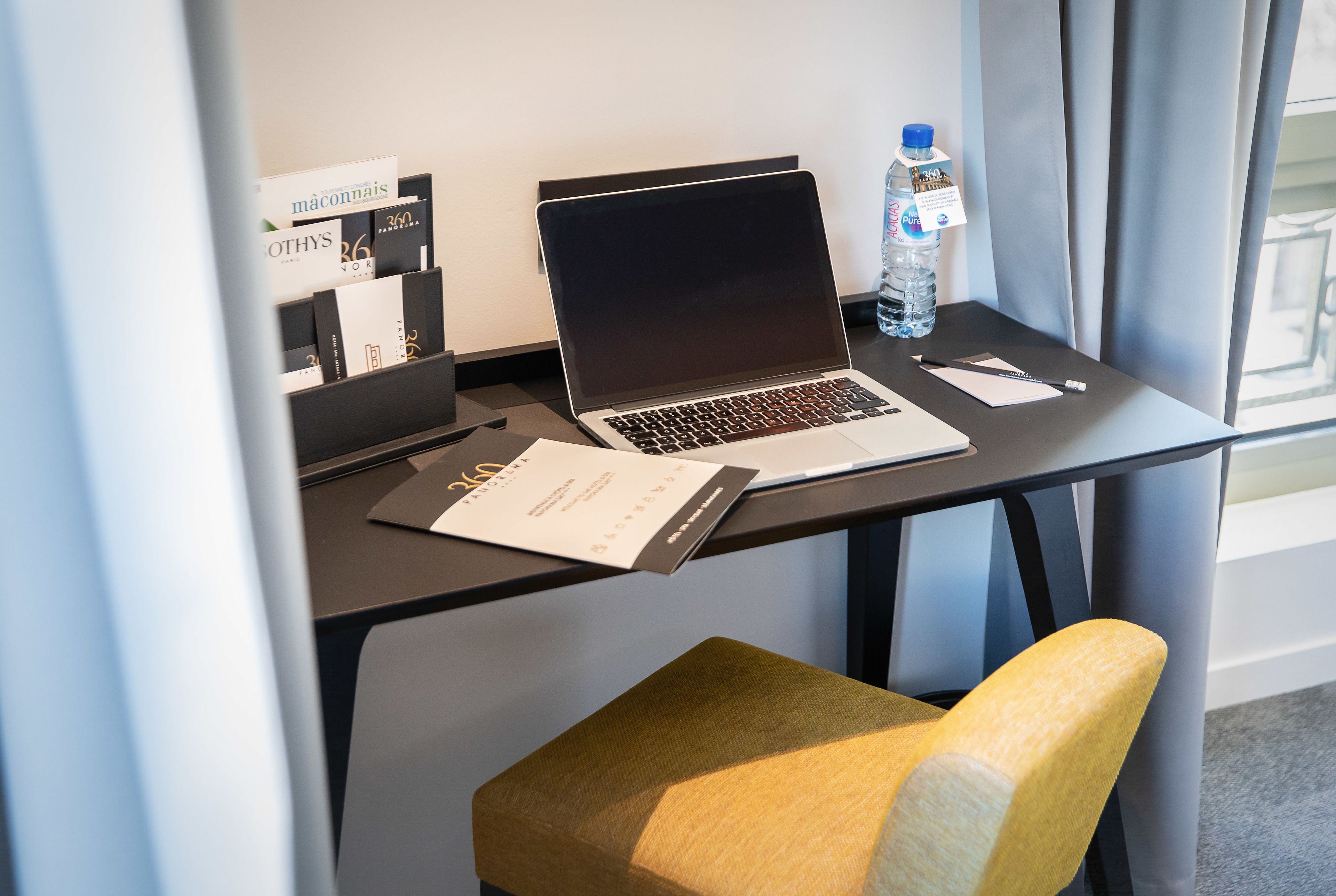 A hotel room with a desk, laptop, water bottle, and a chair.