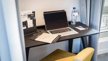 A hotel room with a desk, laptop, water bottle, and a chair.