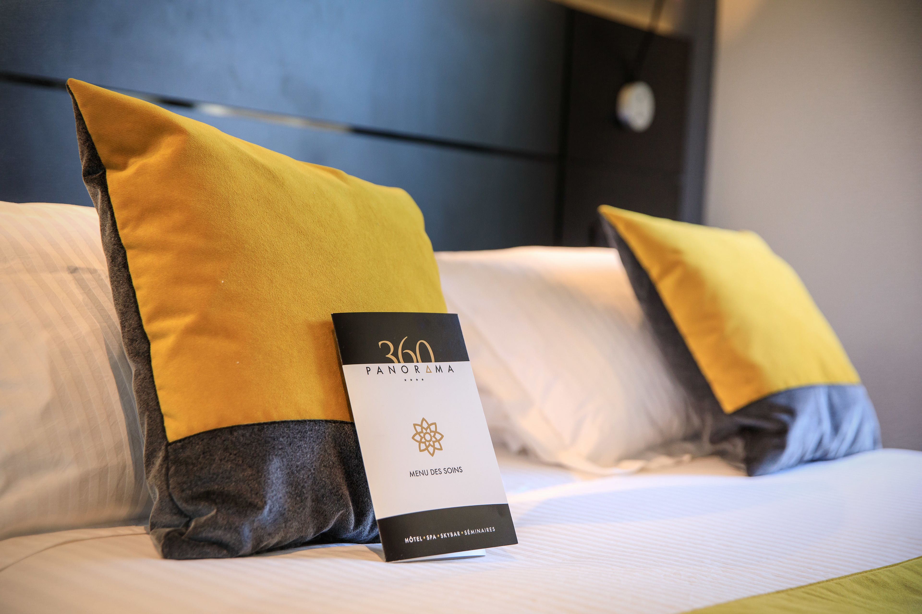 A hotel bed with two pillows, one yellow and one gray, and a menu card labeled "360 PANORAMA MENU DES SOINS HOTEL SPA SKYBAS SEMINAIRES".